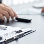 This image shows a professional office setting focused on automotive or insurance business. In the foreground, there's a clipboard with documents and a silver pen lying on top. Next to the clipboard sits a small golden-yellow toy or model car. In the background, two people are working - one person's hands are visible using a black calculator, while another person's hands can be seen near what appears to be a computer keyboard. The scene is set on a clean white desk surface, suggesting this might be an insurance office, car dealership, or financial services location where automotive-related paperwork and calculations are being processed. The lighting is bright and professional, creating a business-like atmosphere.