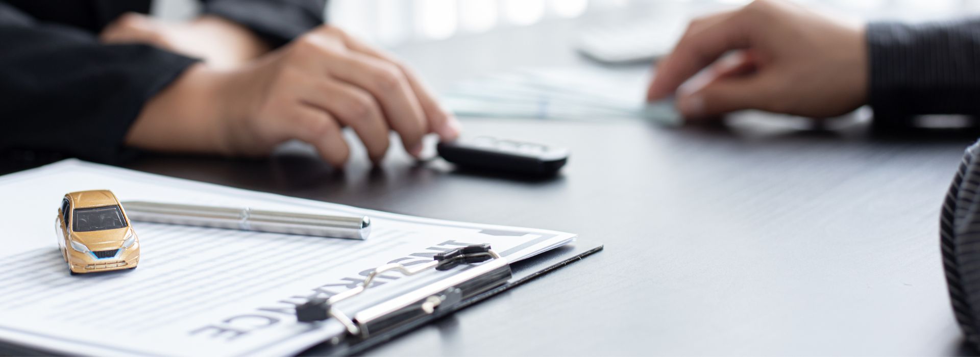 This image shows a professional office setting focused on automotive or insurance business. In the foreground, there's a clipboard with documents and a silver pen lying on top. Next to the clipboard sits a small golden-yellow toy or model car. In the background, two people are working - one person's hands are visible using a black calculator, while another person's hands can be seen near what appears to be a computer keyboard. The scene is set on a clean white desk surface, suggesting this might be an insurance office, car dealership, or financial services location where automotive-related paperwork and calculations are being processed. The lighting is bright and professional, creating a business-like atmosphere.