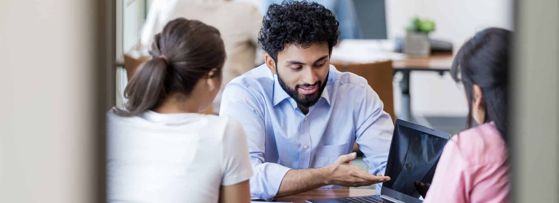 The image shows three people in a business meeting or consultation. In the center, a man with dark curly hair and a beard wearing a light blue shirt is pointing at or explaining something on a laptop. He's seated at a table with two women - one with her back partially to the camera wearing a white top, and another person wearing pink visible on the right side. The setting appears to be a bright office space with natural lighting coming through windows in the background.
