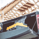A construction worker wearing a yellow hard hat, plaid shirt, and work gloves loads a yellow chainsaw into the bed of a black pickup truck. The worker has a tool belt around his waist and is positioned at the truck's tailgate. In the background, there's a wooden house frame under construction with exposed roof trusses and rafters against a cloudy sky, indicating an active residential building site.