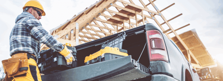 A construction worker wearing a yellow hard hat, plaid shirt, and work gloves loads a yellow chainsaw into the bed of a black pickup truck. The worker has a tool belt around his waist and is positioned at the truck's tailgate. In the background, there's a wooden house frame under construction with exposed roof trusses and rafters against a cloudy sky, indicating an active residential building site.