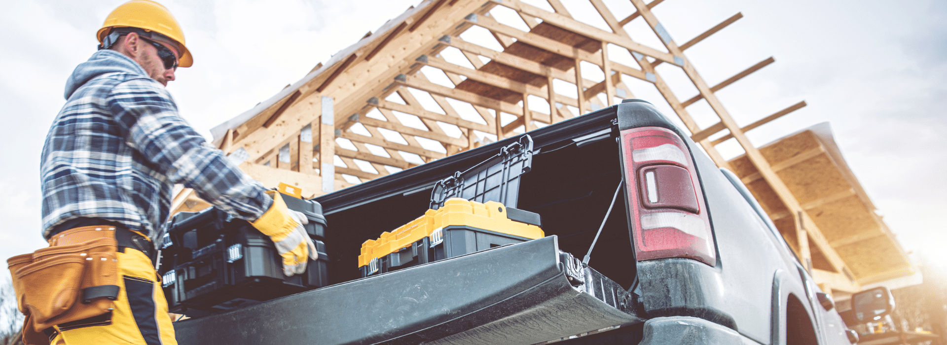 A construction worker wearing a yellow hard hat, plaid shirt, and work gloves loads a yellow chainsaw into the bed of a black pickup truck. The worker has a tool belt around his waist and is positioned at the truck's tailgate. In the background, there's a wooden house frame under construction with exposed roof trusses and rafters against a cloudy sky, indicating an active residential building site.