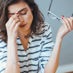 A woman sitting at a desk looks stressed, rubbing her eyes while holding her glasses in one hand. She is surrounded by an open notebook, a glass of water, a tablet, and a laptop, suggesting she is overwhelmed with work or financial concerns in a home office setting.