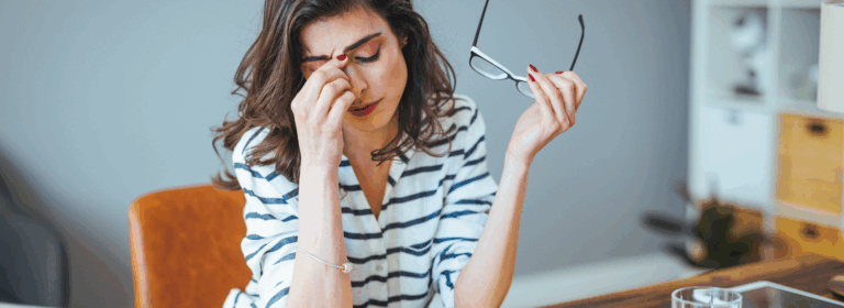 A woman sitting at a desk looks stressed, rubbing her eyes while holding her glasses in one hand. She is surrounded by an open notebook, a glass of water, a tablet, and a laptop, suggesting she is overwhelmed with work or financial concerns in a home office setting.