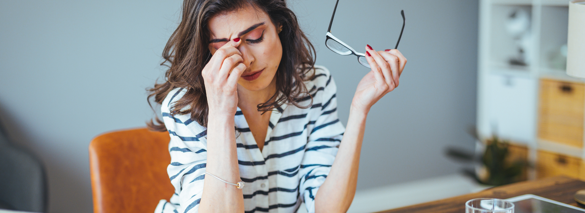 A woman sitting at a desk looks stressed, rubbing her eyes while holding her glasses in one hand. She is surrounded by an open notebook, a glass of water, a tablet, and a laptop, suggesting she is overwhelmed with work or financial concerns in a home office setting.